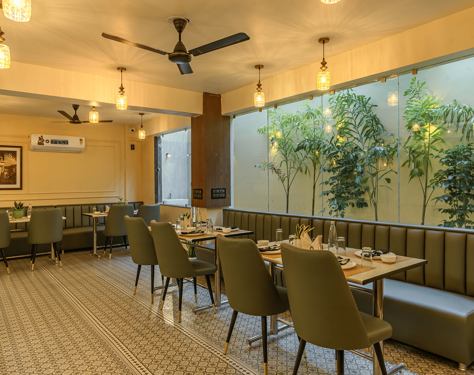 Restaurant at Perfectstayz Premium Lavender, Varanasi with olive green chairs and banquettes, pendant lamps, patterned floor tiles, and a glass wall with plants.