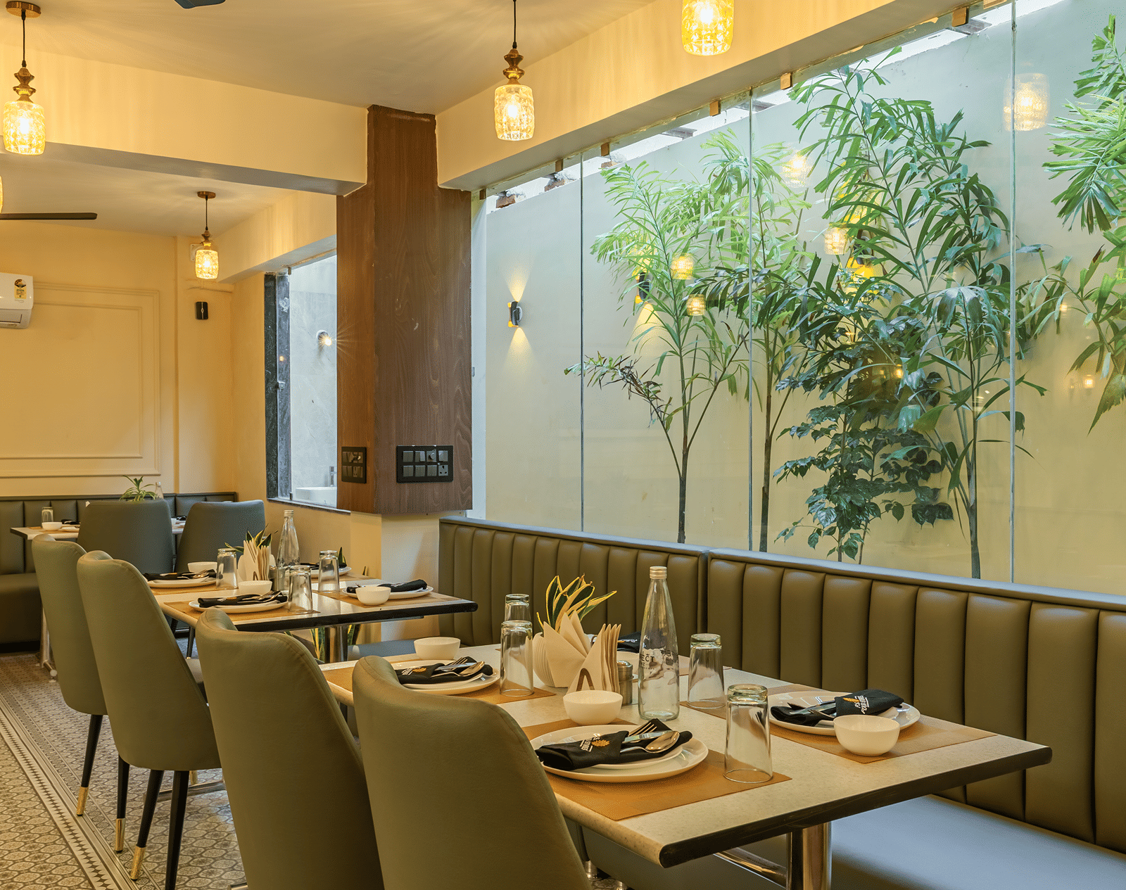 Set dining table at the restaurant of Perfectstayz Premium Lavender, Varanasi with plates, glasses, and a water bottle beside green banquette seating and plants.