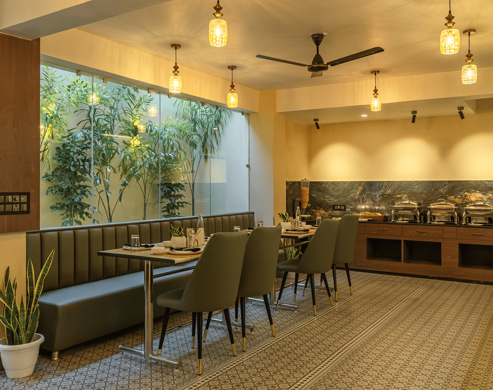 Restaurant at Perfectstayz Premium Lavender, Varanasi with a buffet counter of 4 chafing dishes, green banquette seating, pendant lamps, and a planted glass wall.