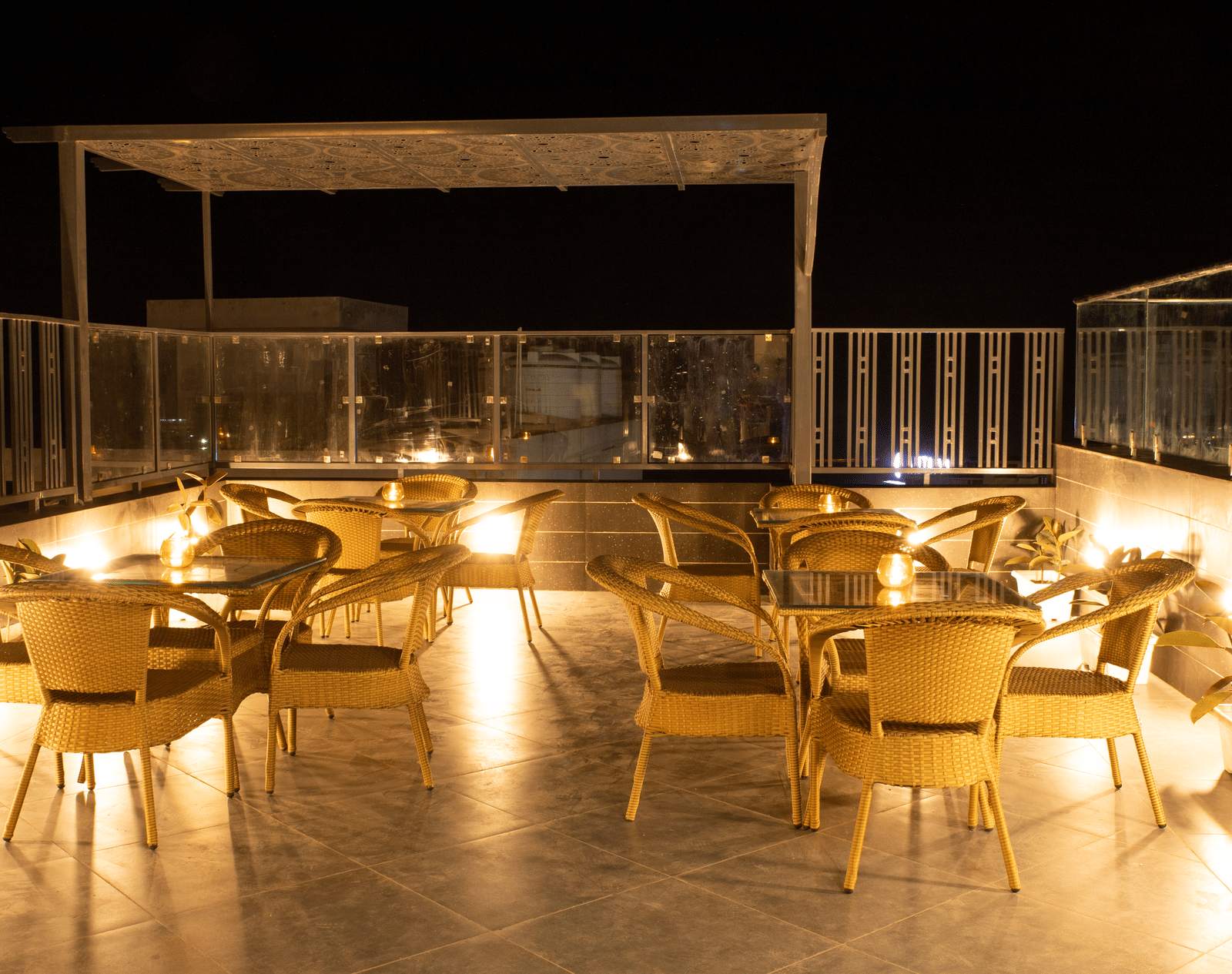 An outdoor rooftop cafe at Perfectstayz Premium Laxmi Heritage with woven chairs and tables on a patio, warmly lit under a dark sky, enclosed by a glass railing.