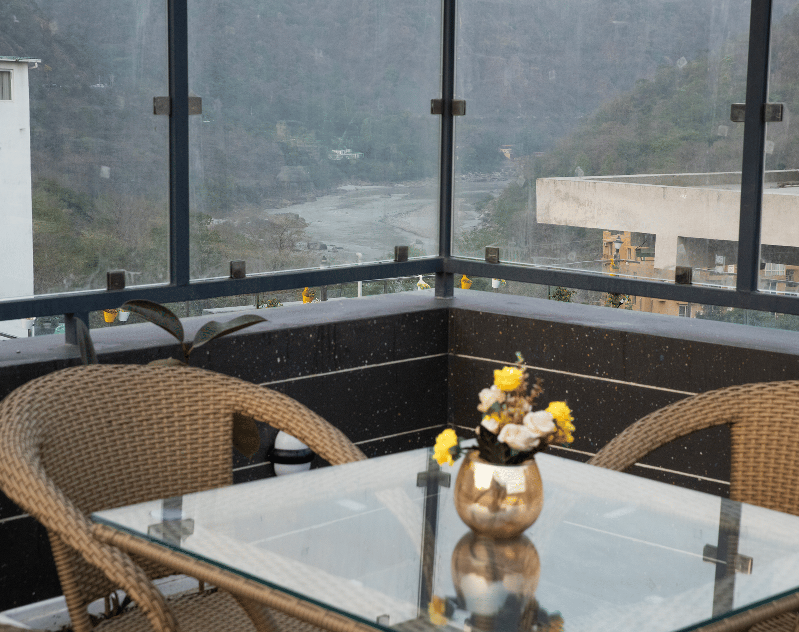 An outdoor dining area at Perfectstayz Premium Laxmi Heritage with wicker chairs, a glass-top table, a small bouquet, and views of a river and mountains.