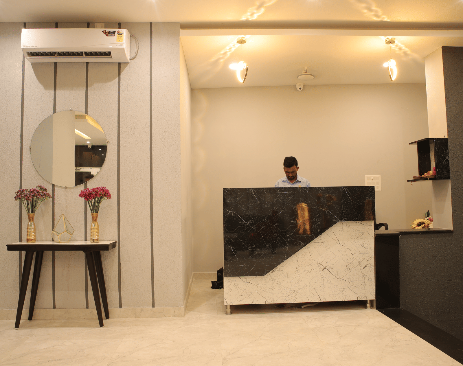 A man standing behind a reception desk at Perfectstayz Premium Laxmi Heritage, with light walls, ceiling lights, a mirror, flower vases, and an air conditioner above.