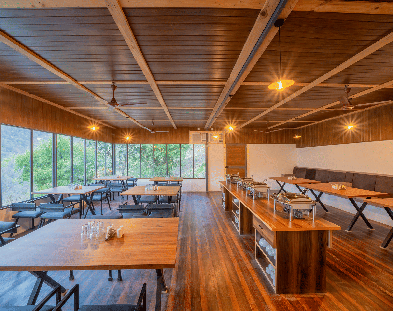 The dining area at Perfect Stayz The Jungle Resort features wooden tables, bench seating, a buffet counter, and windows looking out at nature.