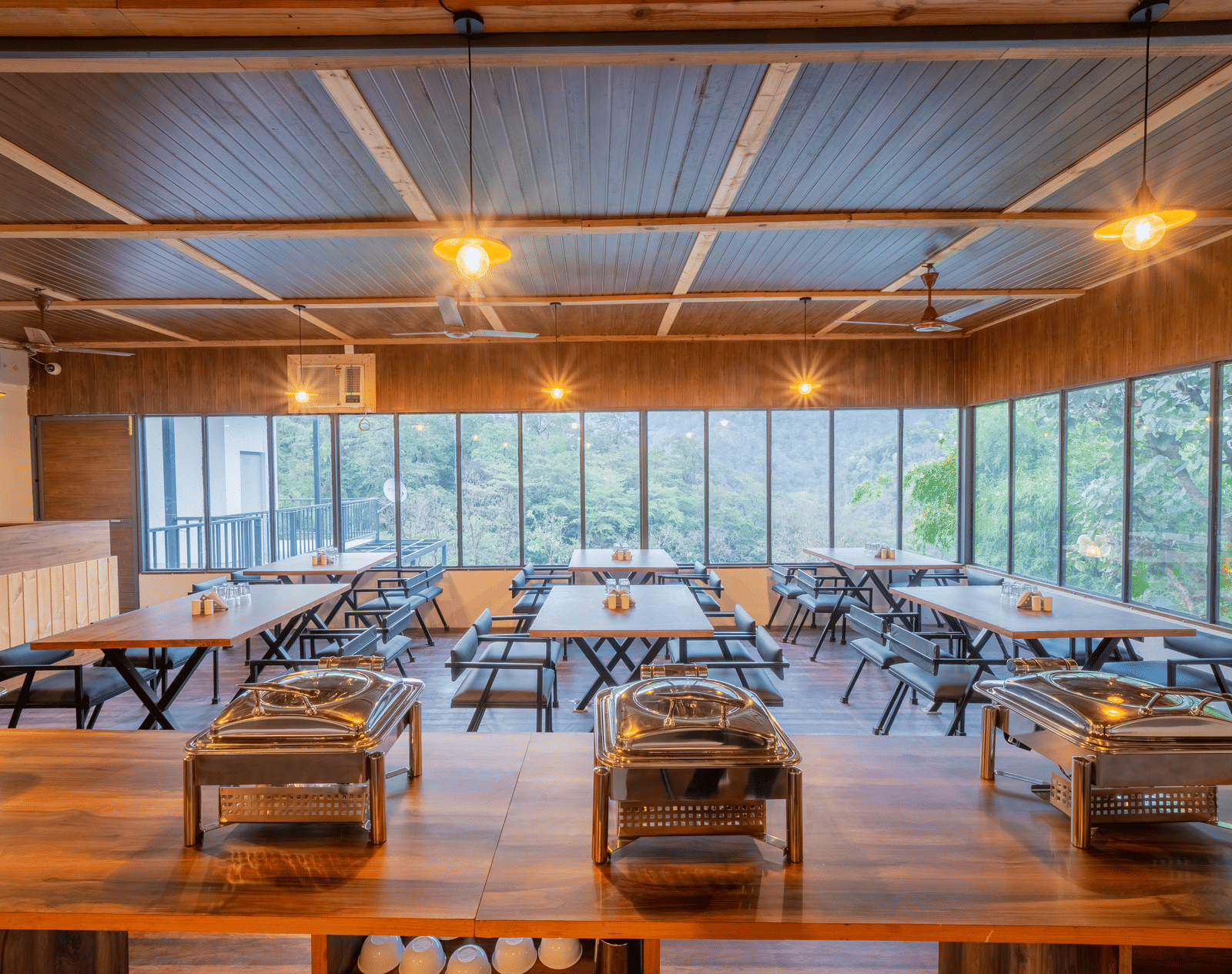 The dining hall at Perfect Stayz The Jungle Resort has long tables, chairs, chafing dishes, and windows overlooking trees.