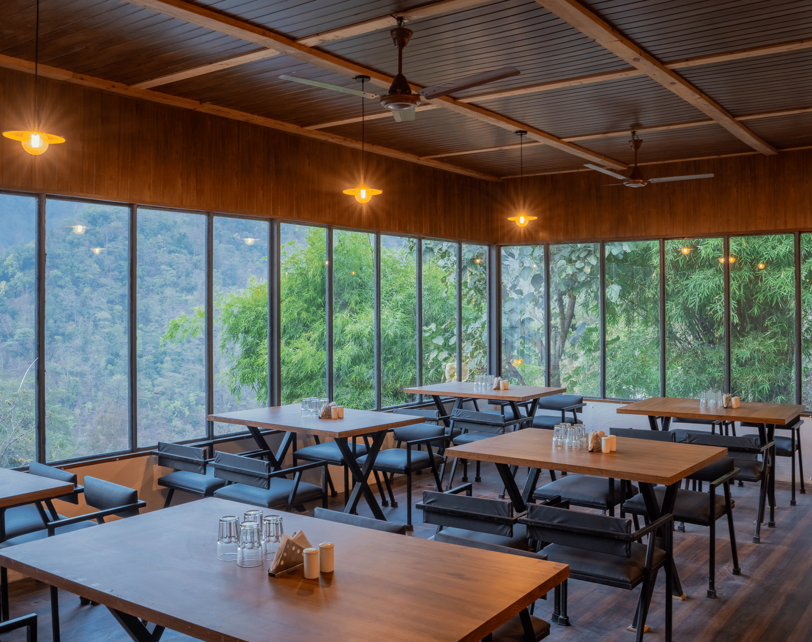 A restaurant area at Perfect Stayz The Jungle Resort has tables, chairs, and glass walls overlooking nature.
