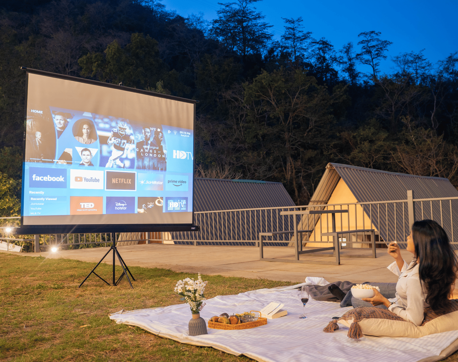 A person sits on a picnic blanket watching a film projected onto a screen at Perfect Stayz The Jungle Resort.