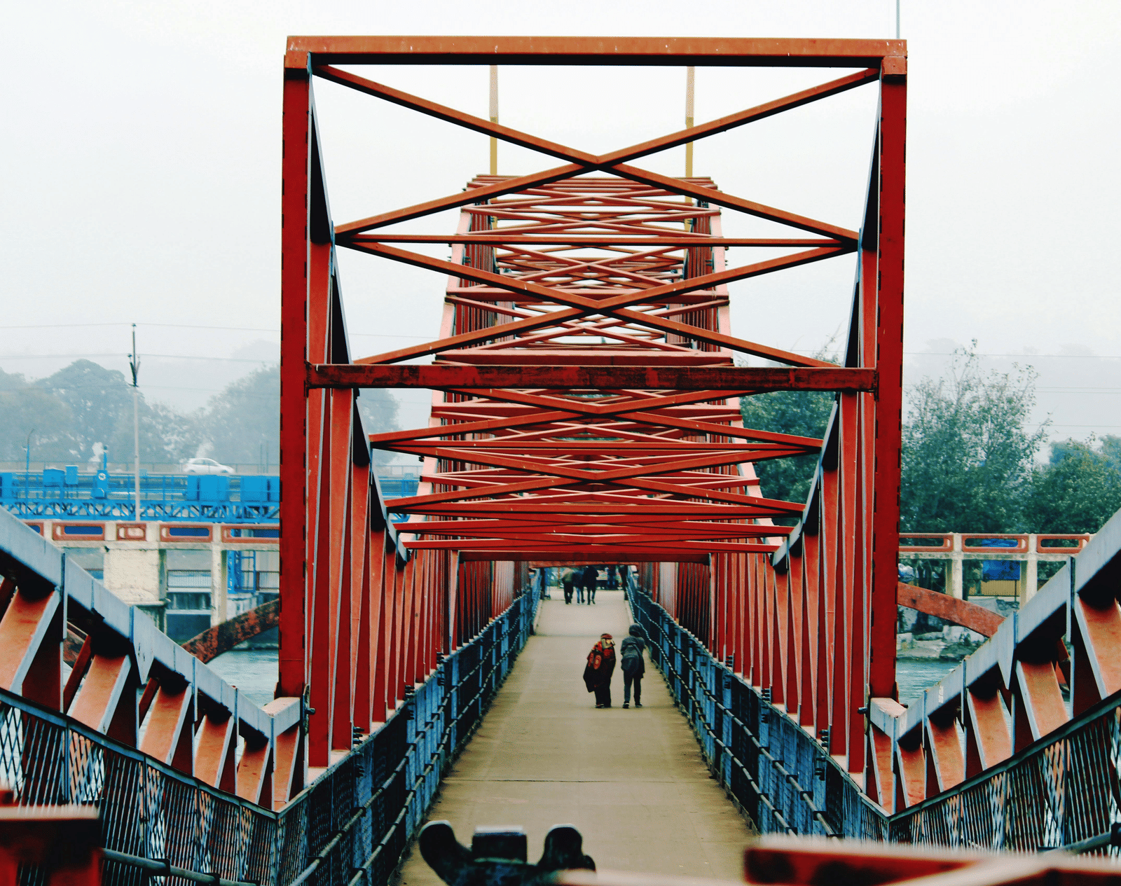 Pedestrian bridge with striking red metal framework in Haridwar, India, featuring people walking across on a misty day.