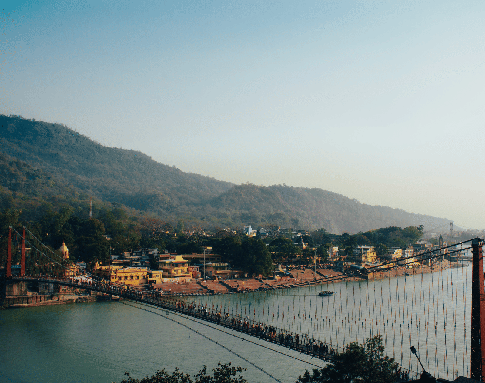 A long suspension bridge spanning a river in a valley, with hills and a town visible in the background