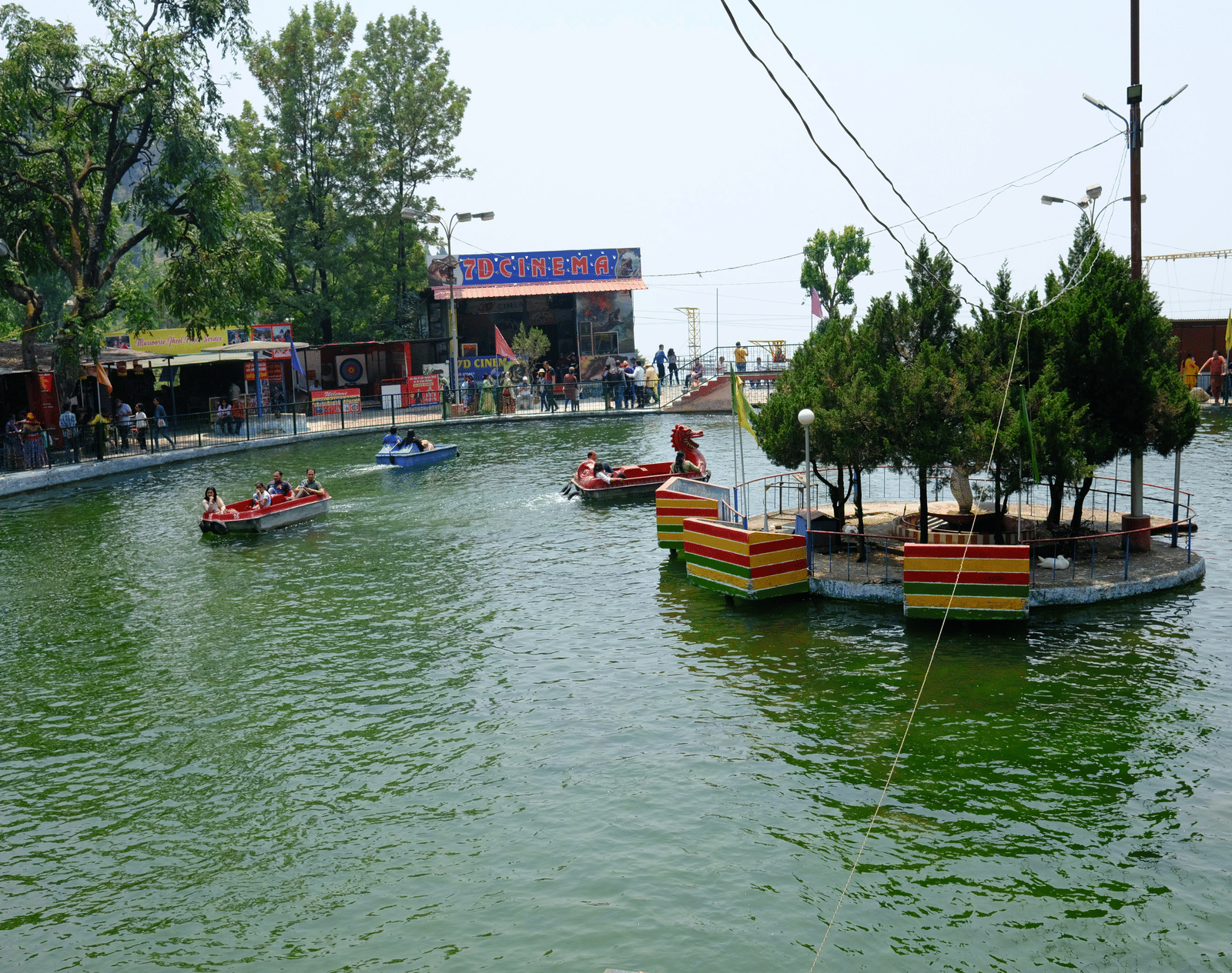 A boating lake with colourful paddle boats and a small tree-covered island surrounded by visitors enjoying the water.