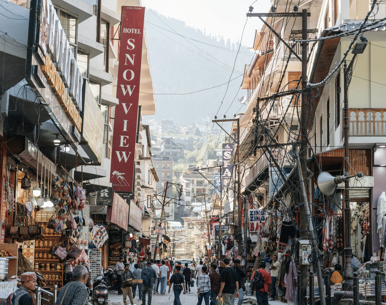 A busy street with shops, hanging signboards, electric poles, and people walking between buildings in a market area with hills in the background.