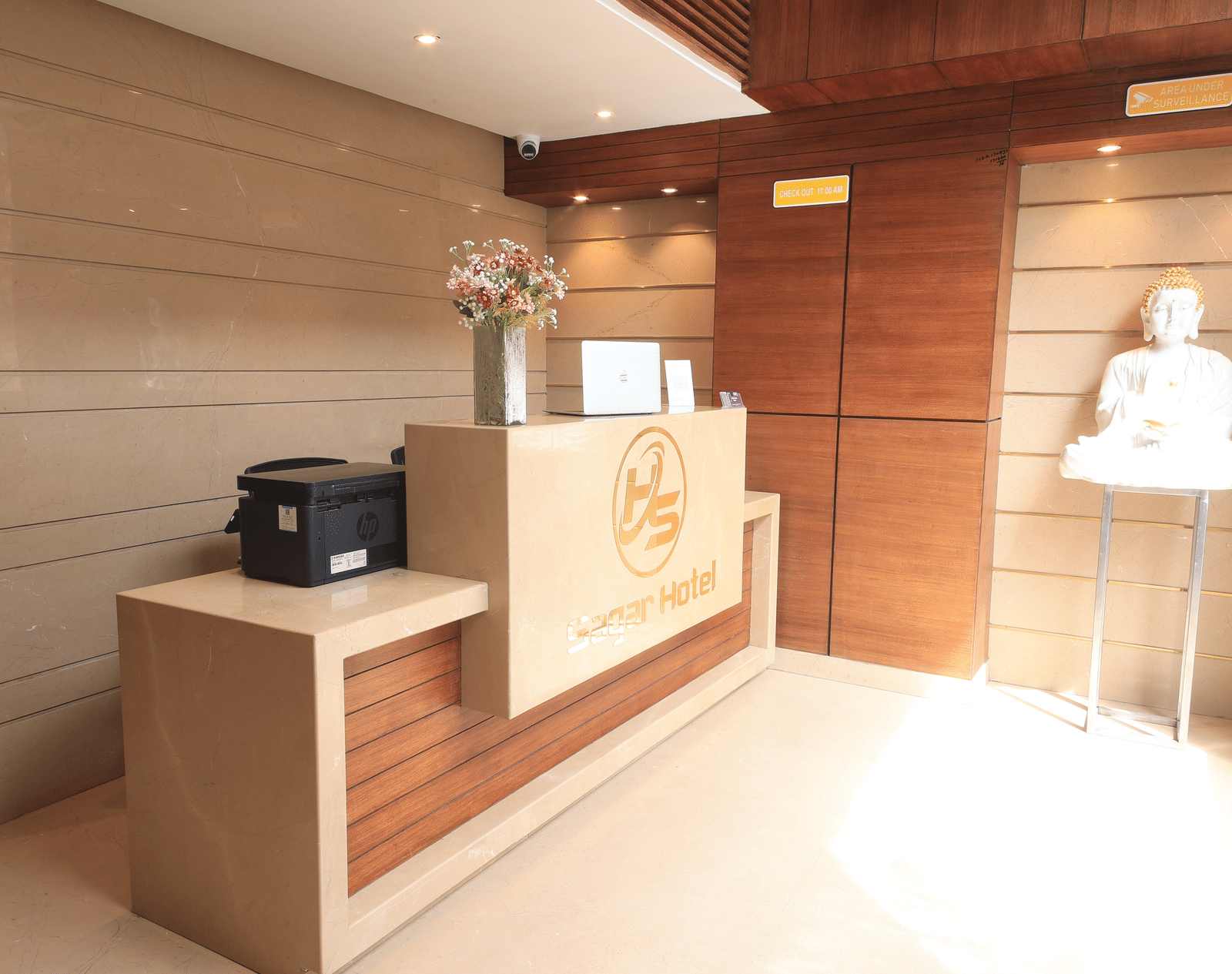 A hotel reception area, featuring a reception desk with a logo, a printer, and a statue on a stand | Perfectstayz Value Sagar