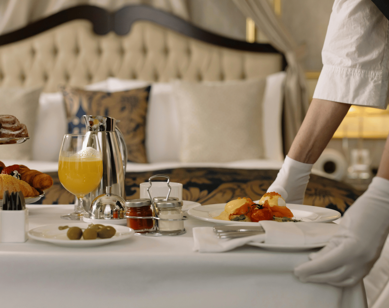 Two hands placing a bowl of fruits on a table filled with eateries near a bed.