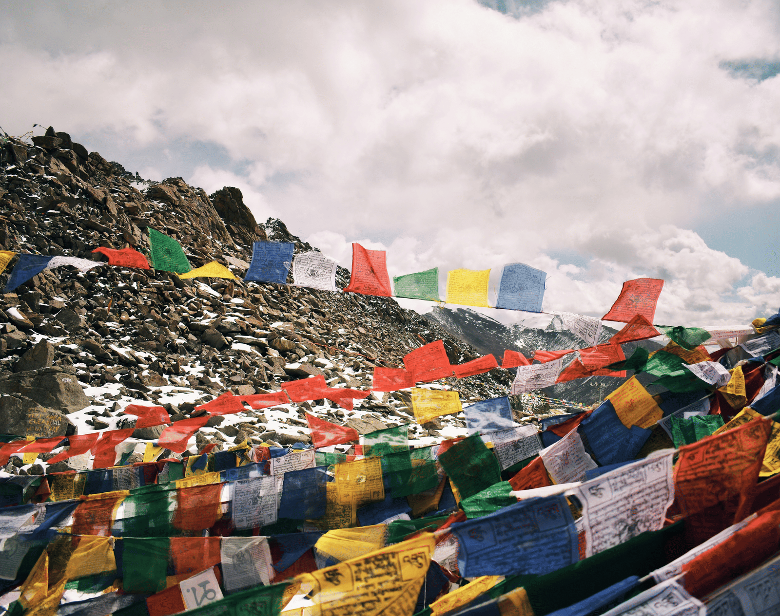 A mountain landscape with a rocky peak, covered in colourful Buddhist prayer flags waving under a cloudy sky