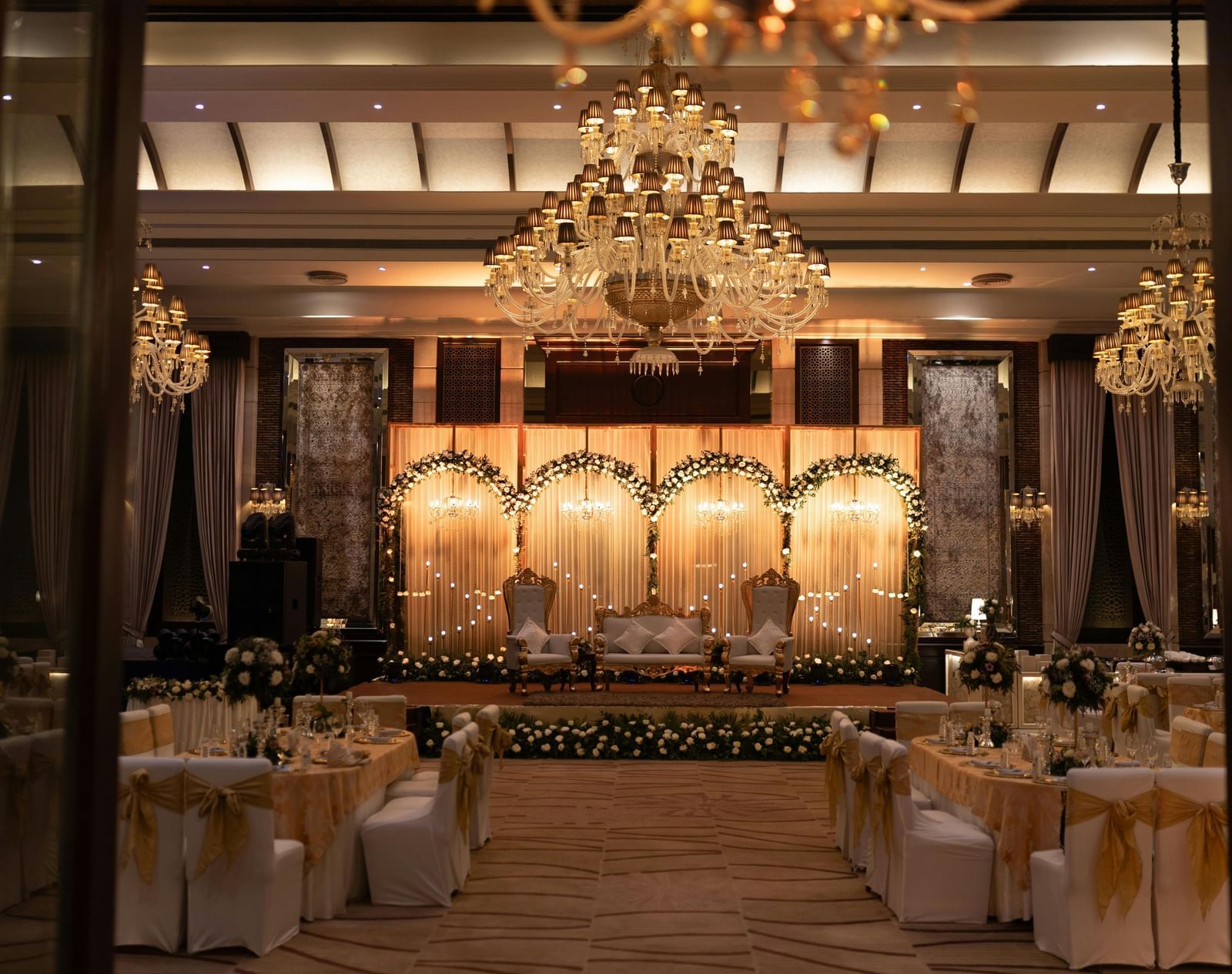A grand banquet hall with chandeliers, golden decor, and elegantly arranged round tables.
