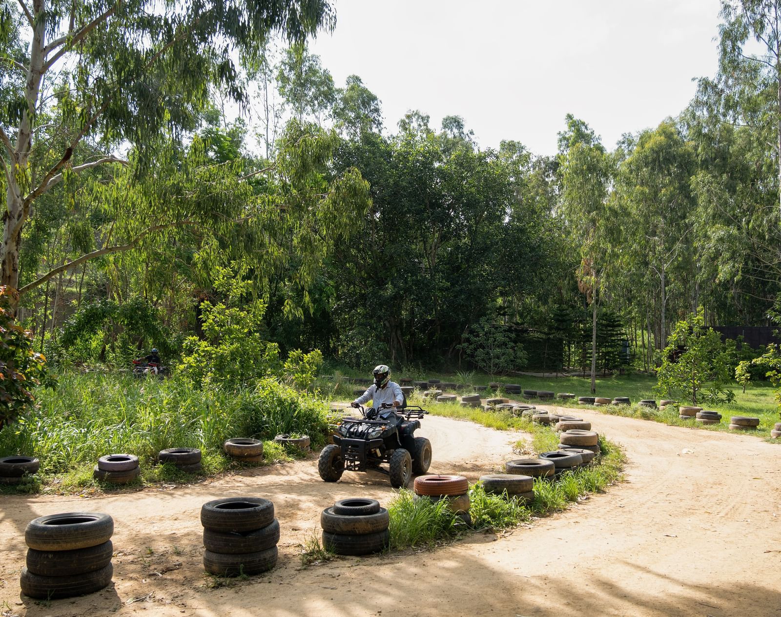 A winding dirt path, lined with tires, leads through a scenic forested area under a bright sky | RD's Nature Retreat, Bangalore