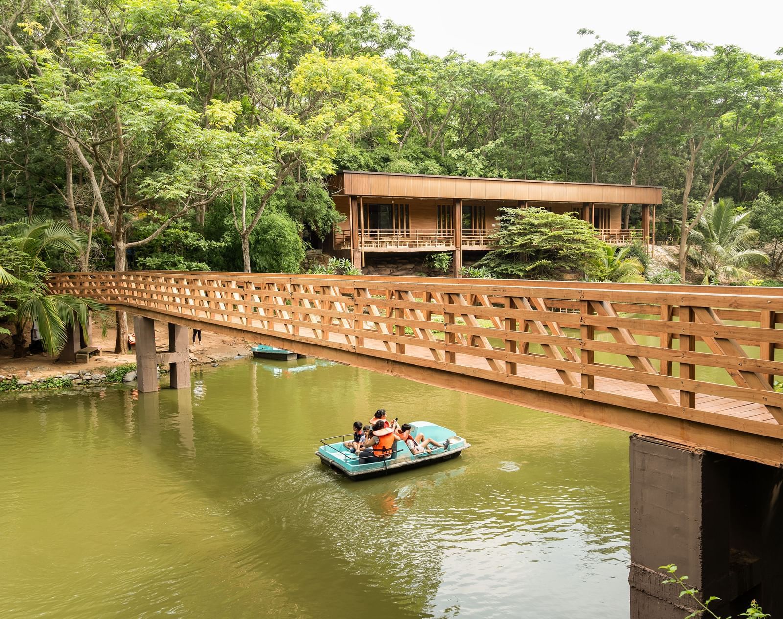 A wooden bridge spans across a calm body of water where a small boat is visible, surrounded by abundant green foliage | Lake View Restaurant