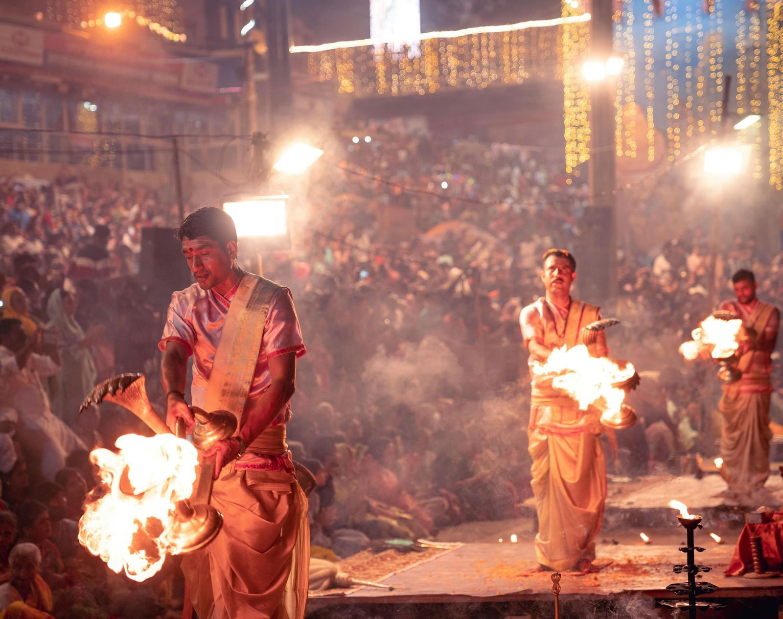 Several priests performing Ganga Aarti with a huge crowd of devotees gathered behind them.