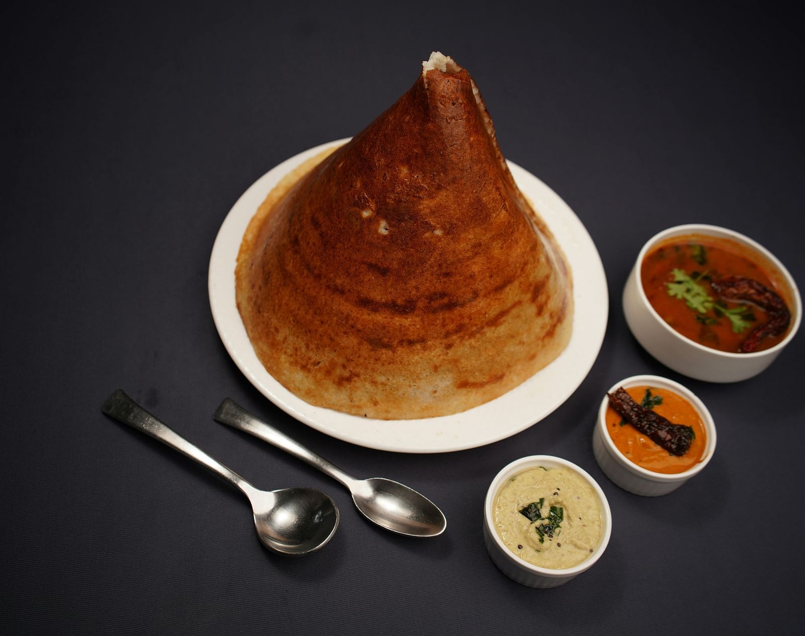 A golden dosa on a white plate, accompanied by three small bowls of colourful chutneys and sambar, with two spoons resting beside it.