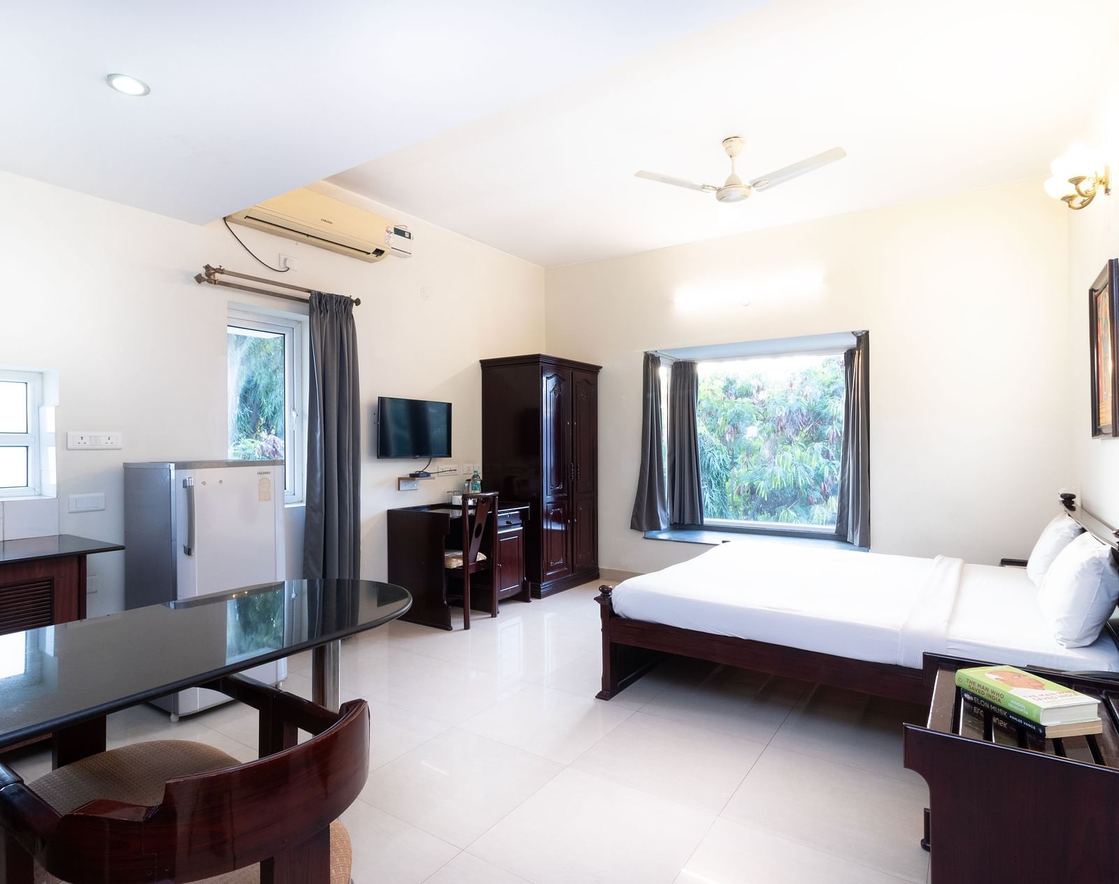 A spacious room offering a double bed with white linens, a dining table with chairs, a television, a refrigerator, and a window providing natural light at Sanctum Suites Indiranagar, Bangalore.