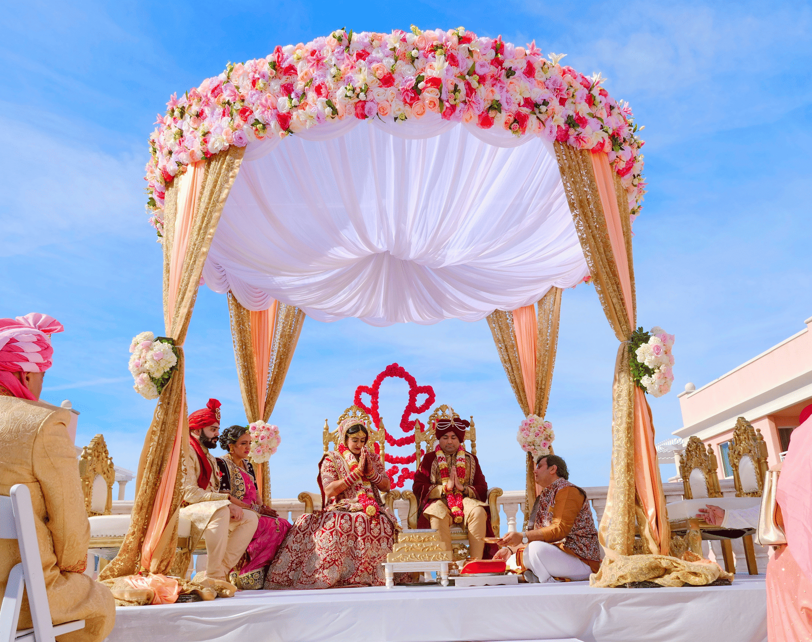The wedding ceremony is underway in a decorative outdoor mandap with a pink and white floral canopy, set against a bright blue sky.