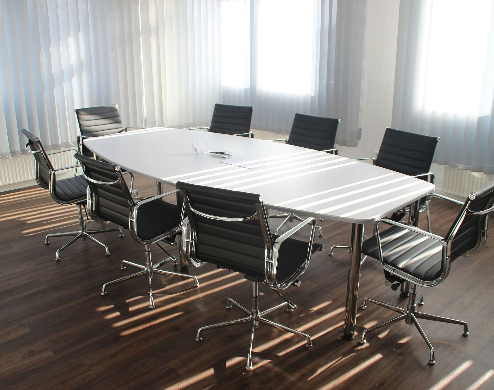 A close up shot of a brightly lit boardroom featuring a white long table along with office chairs surrounding it.
