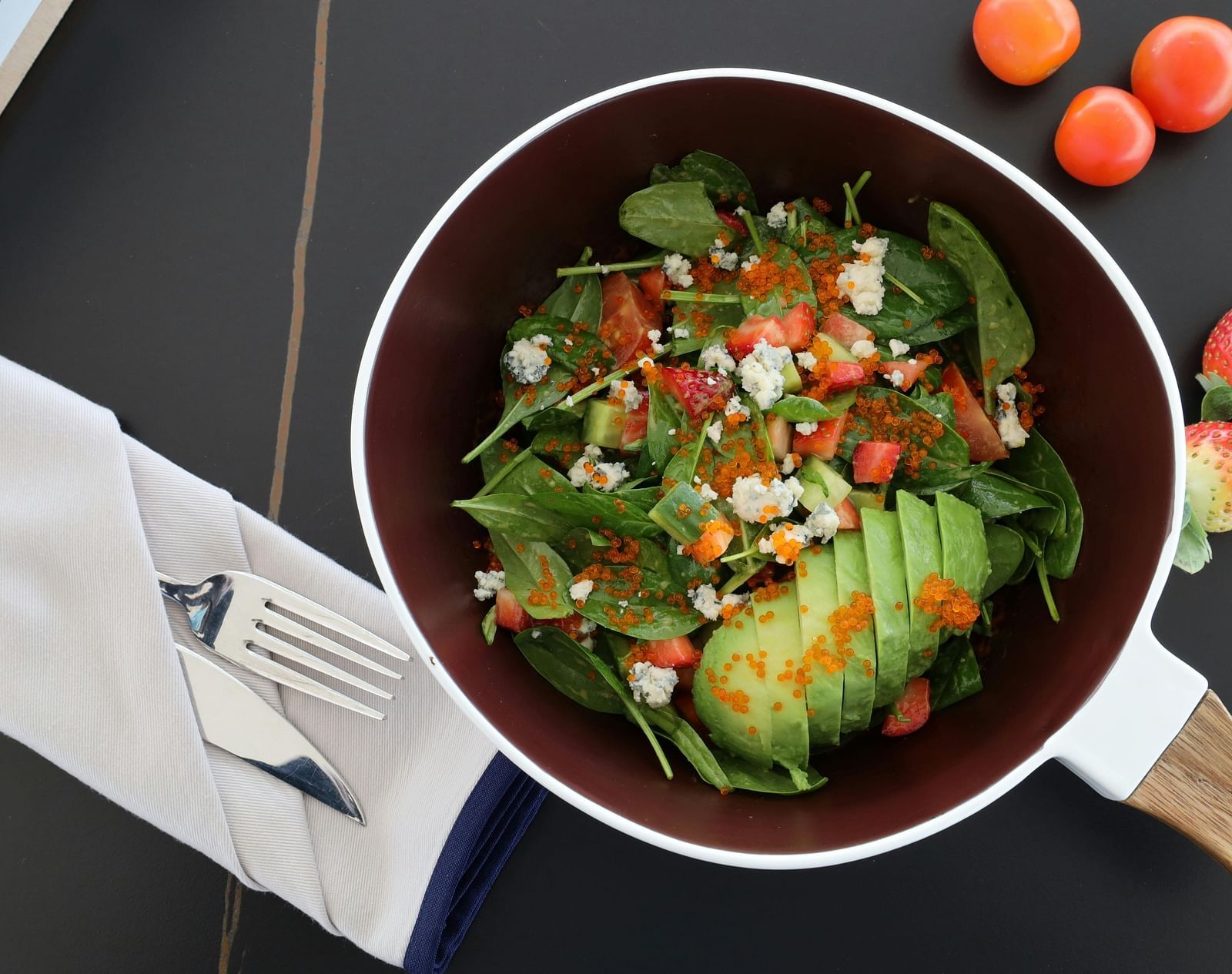 A top-shot of a spinach salad with avocado served in a pan next to cutlery.