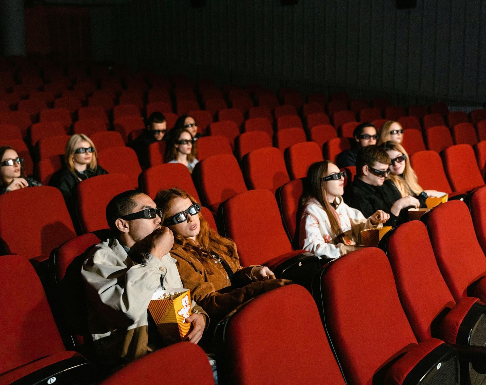 People wearing masks are seated in red theater seats, watching a screen in a dark movie theater.