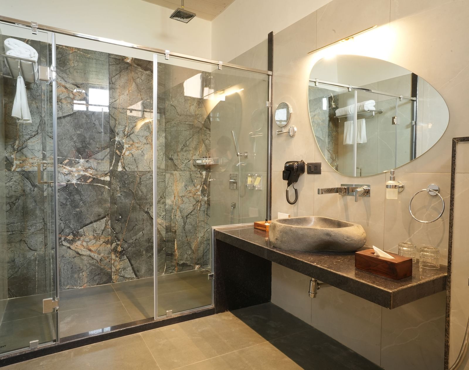 A modern bathroom of Club Suite with Valley View Sitout with a glass-enclosed shower, a unique-shaped mirror, and a sleek vanity area at The Qualia Resort Club & Brewery, Udaipur.