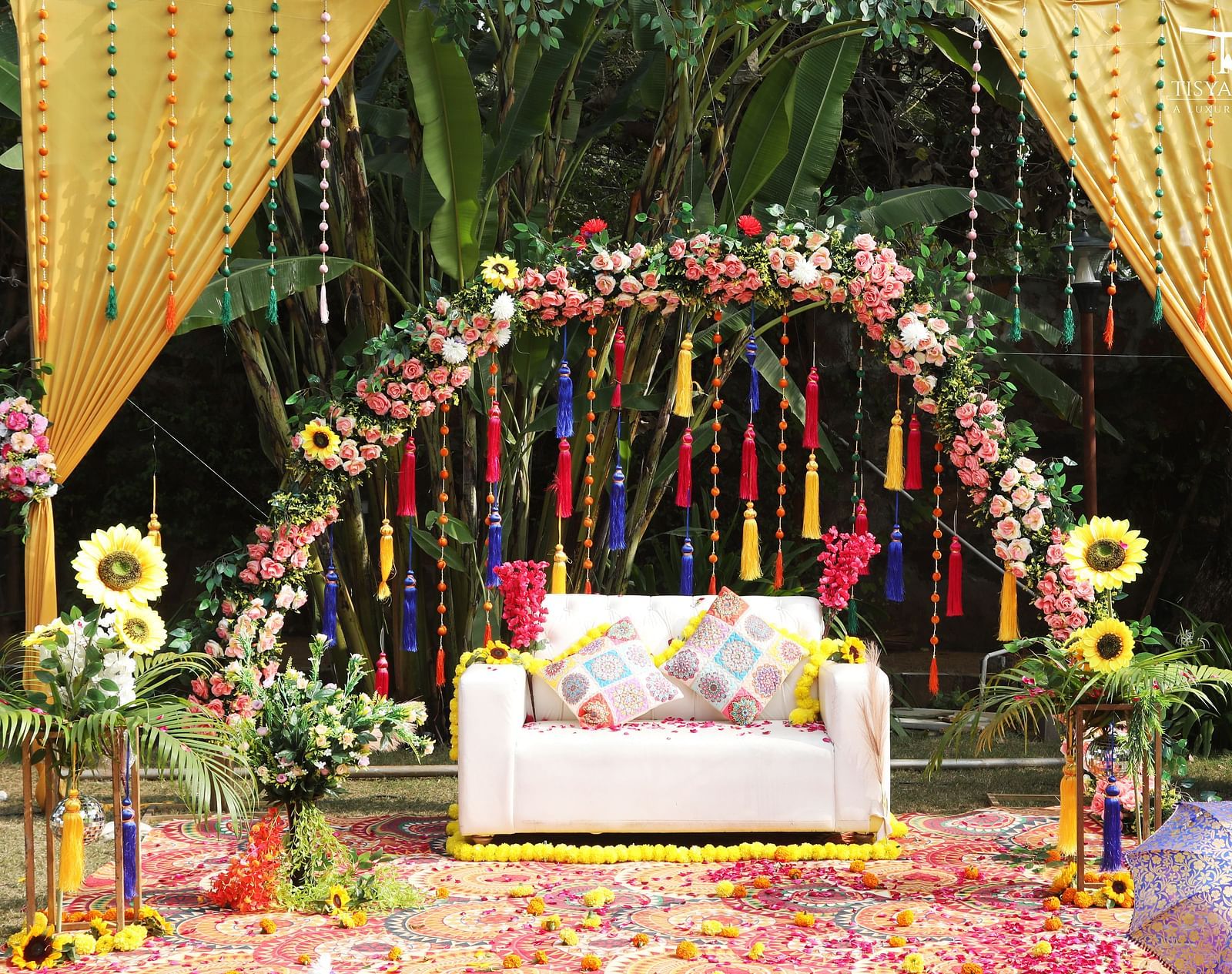 A wedding stage with a white sofa under a floral arch, decorated with hanging tassels and sunflowers at Tisya Farms.