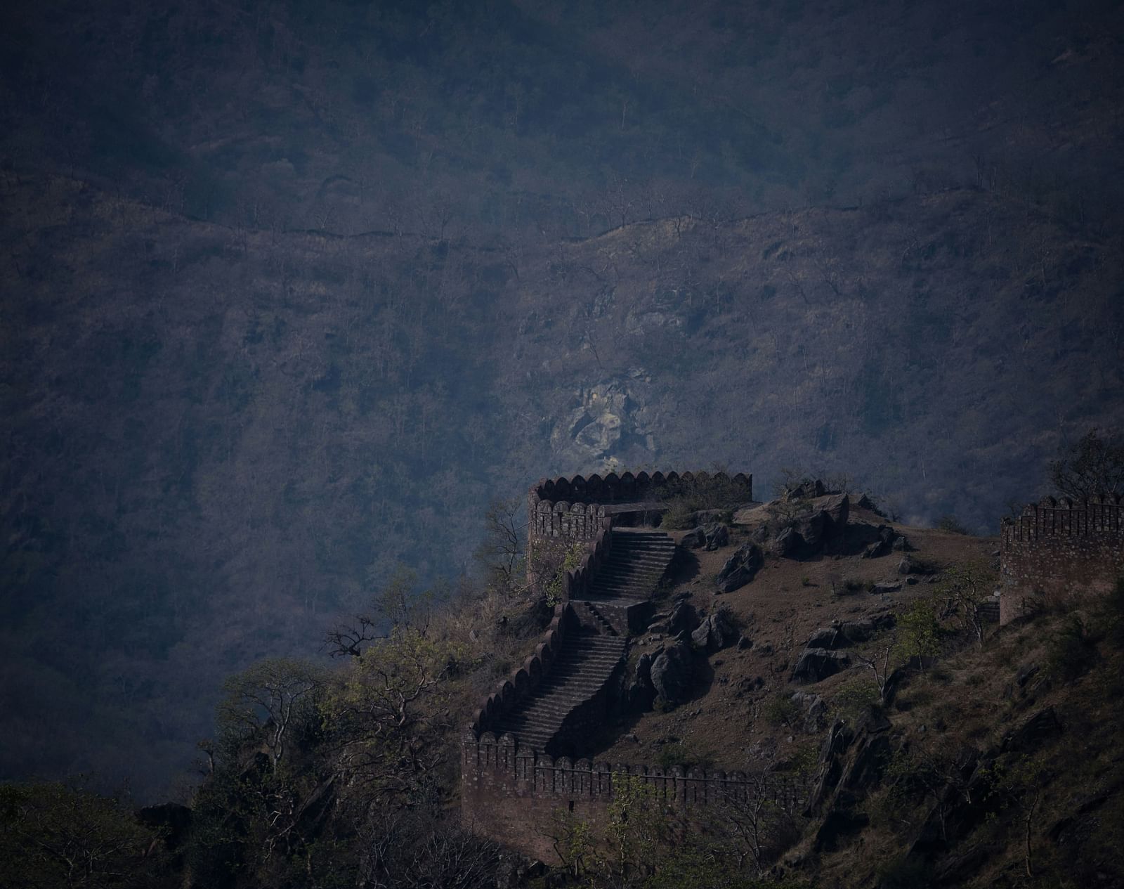 A weathered Kumbhalgarh fort wall with zig-zag stairs and battlements on a rocky hill, against a hazy mountain backdrop - Kumbhalgarh Tourist Attractions