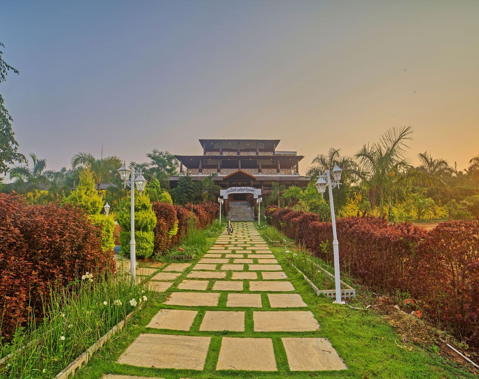 A long pathway of square stepping stones through a vibrant green garden, leading towards a grand, traditional building with multi-tiered roofs under a sunset sky. - Vintage Trails by Ilara