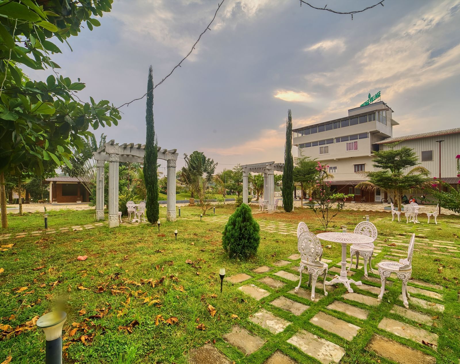 A wide outdoor view of a green lawn with a small table and chairs set, leading towards a modern building under a cloudy sky. - Vintage Trails by Ilara
