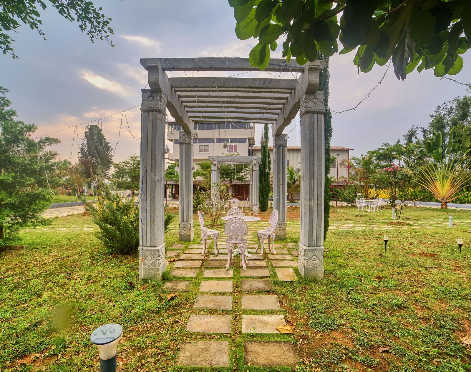 A pathway made of square stepping stones leading towards a modern, open-sided structure with a swing seat, set in a green garden. - Vintage Trails by Ilara