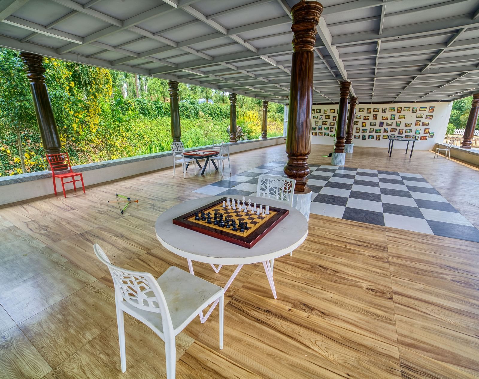A spacious outdoor covered area with a patterned tiled floor, featuring a chess board table and chairs, surrounded by pillars and greenery. - Vintage Trails by Ilara