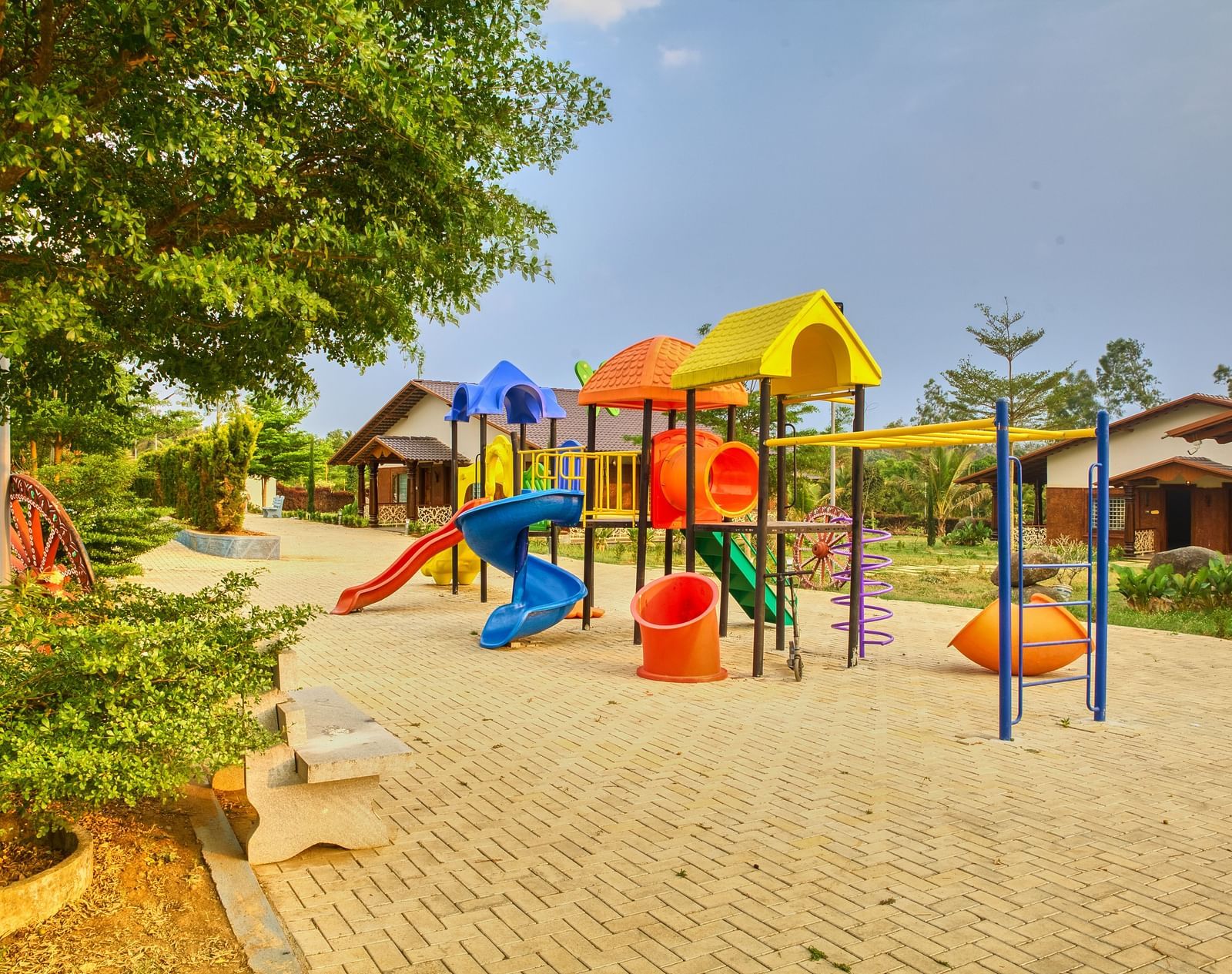 A colourful outdoor children's playground with various slides, climbing frames, and swings on a paved area, surrounded by trees. - Vintage Trails by Ilara