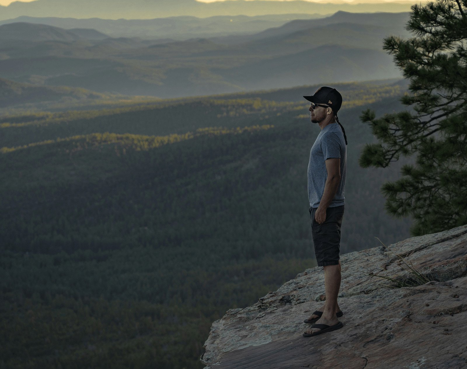 A man in a baseball cap stands on a cliff's edge, gazing at a sprawling forest and mountain range.