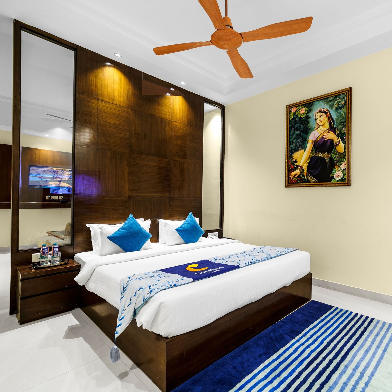 Bedroom at Inde Reva Resorts Rishhikesh with a double bed, blue cushions, ceiling fan, TV, and framed artwork on the wall.