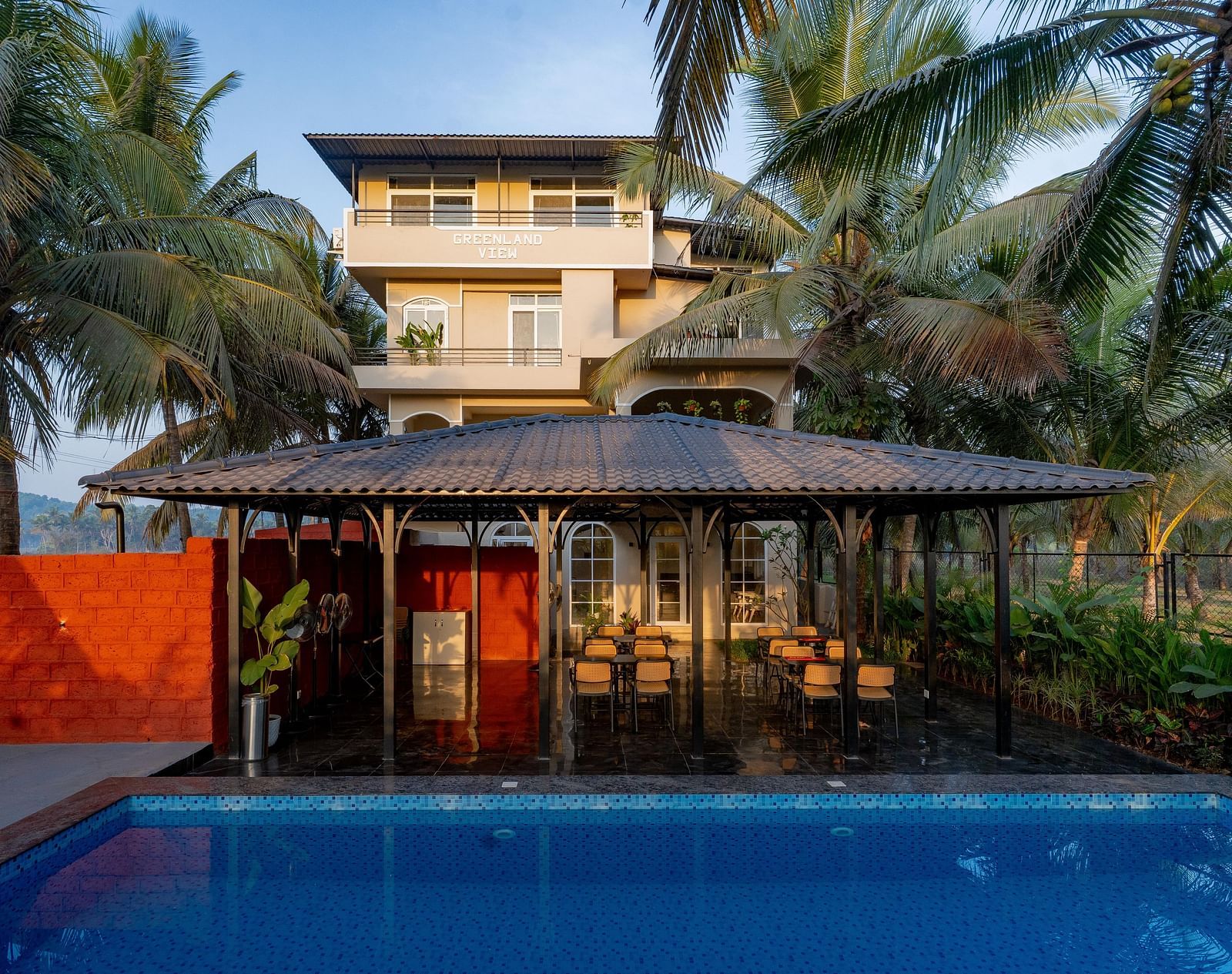A view across a swimming pool towards a covered outdoor seating area with a thatched roof. Palm trees sway gently in the background at Kssemya.