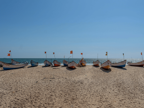 Multiple fishing boats lined along sandy beach under clear sky, with flags attached and sea visible behind the boats.