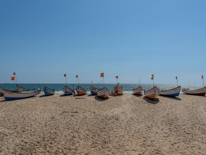 Multiple fishing boats lined along sandy beach under clear sky, with flags attached and sea visible behind the boats.