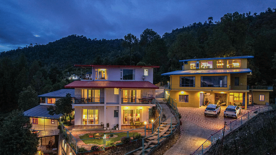 An aerial view of Adrushya Estates, a hill resort in Mukteshwar, on a hillside with lights on and trees around during evening time.