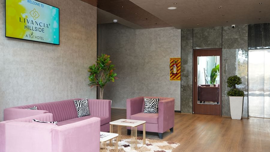 Lounge area with pink sofas, a TV displaying the hotel logo, and decorative plants - TGI Livancia Hillside, JC Nagar, Mysore