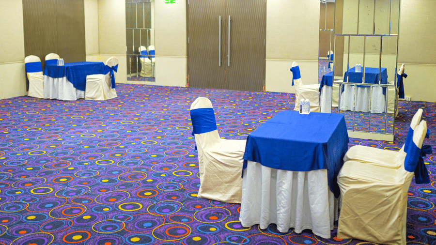 A small banquet room at Ambar Sarovar Portico, Gandhidham displays carpeted floors, patterned ceilings, wooden doors, and tables with matching chairs.