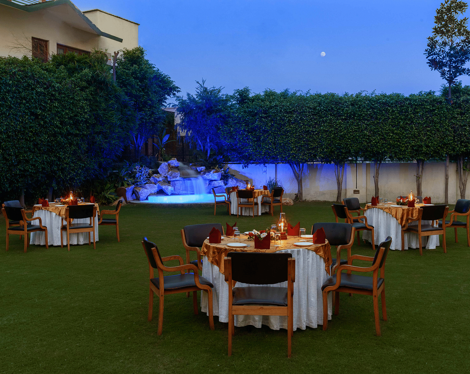 An open lawn with tables and chairs arranged in a circle, each adorned with decorative settings and a small lamp, featuring a lit-up fountain in the corner