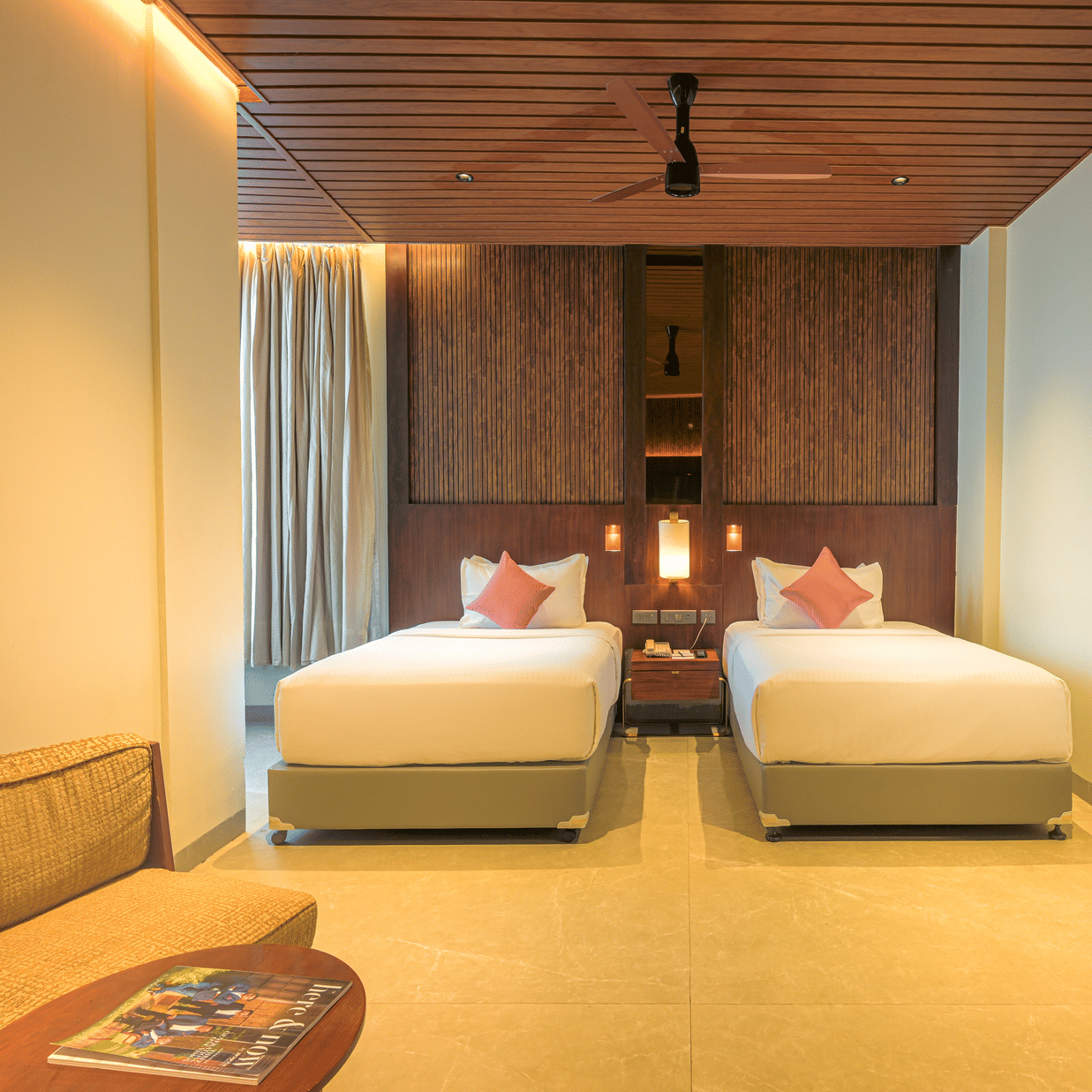 Twin bedroom with two neatly made beds, red accent cushions, wooden ceiling, bedside furnishings and a patterned rug.