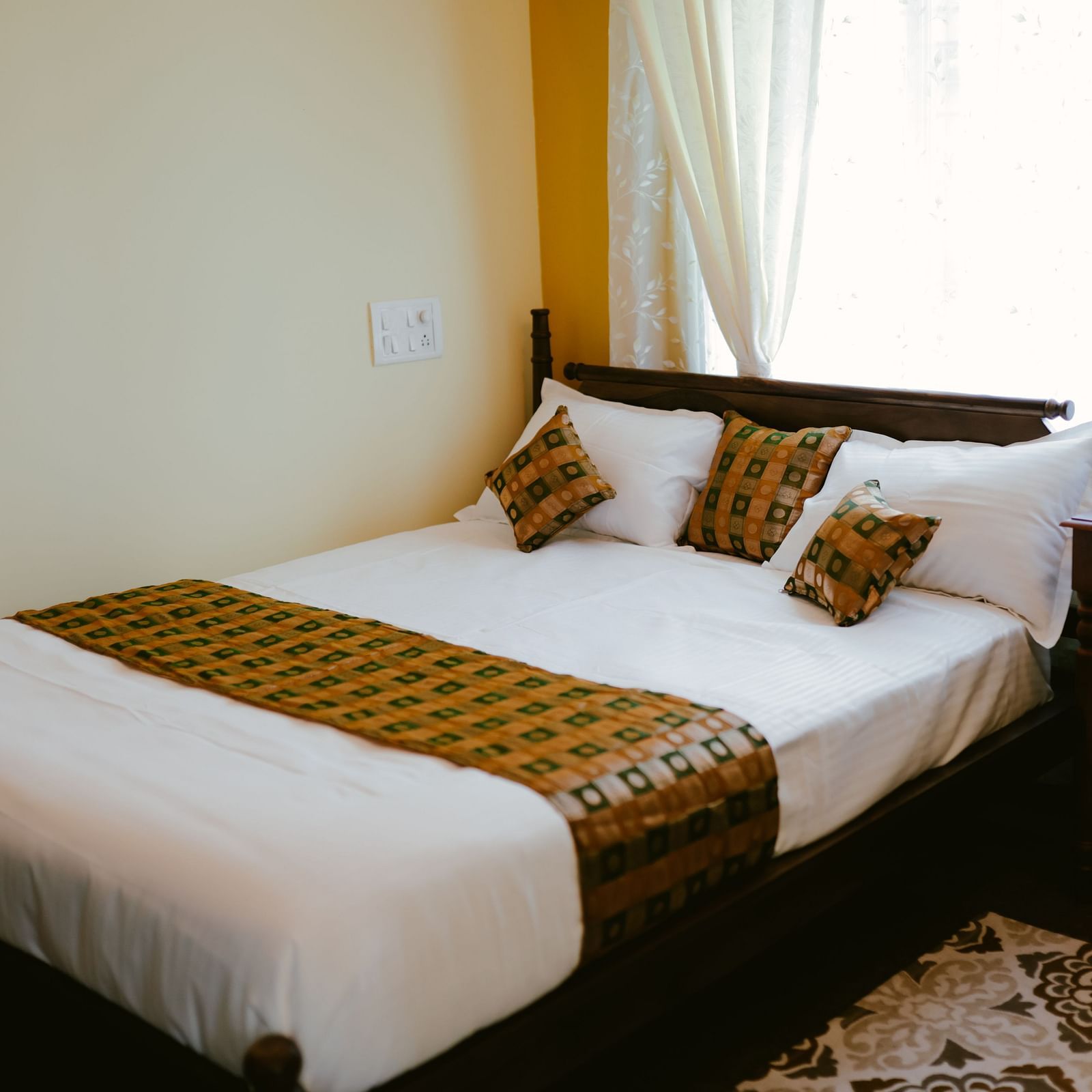 A bedroom with a wooden bed with white linens and patterned throw pillows and a throw blanket, next to a small table | Adhyaya Heritage Reverbed