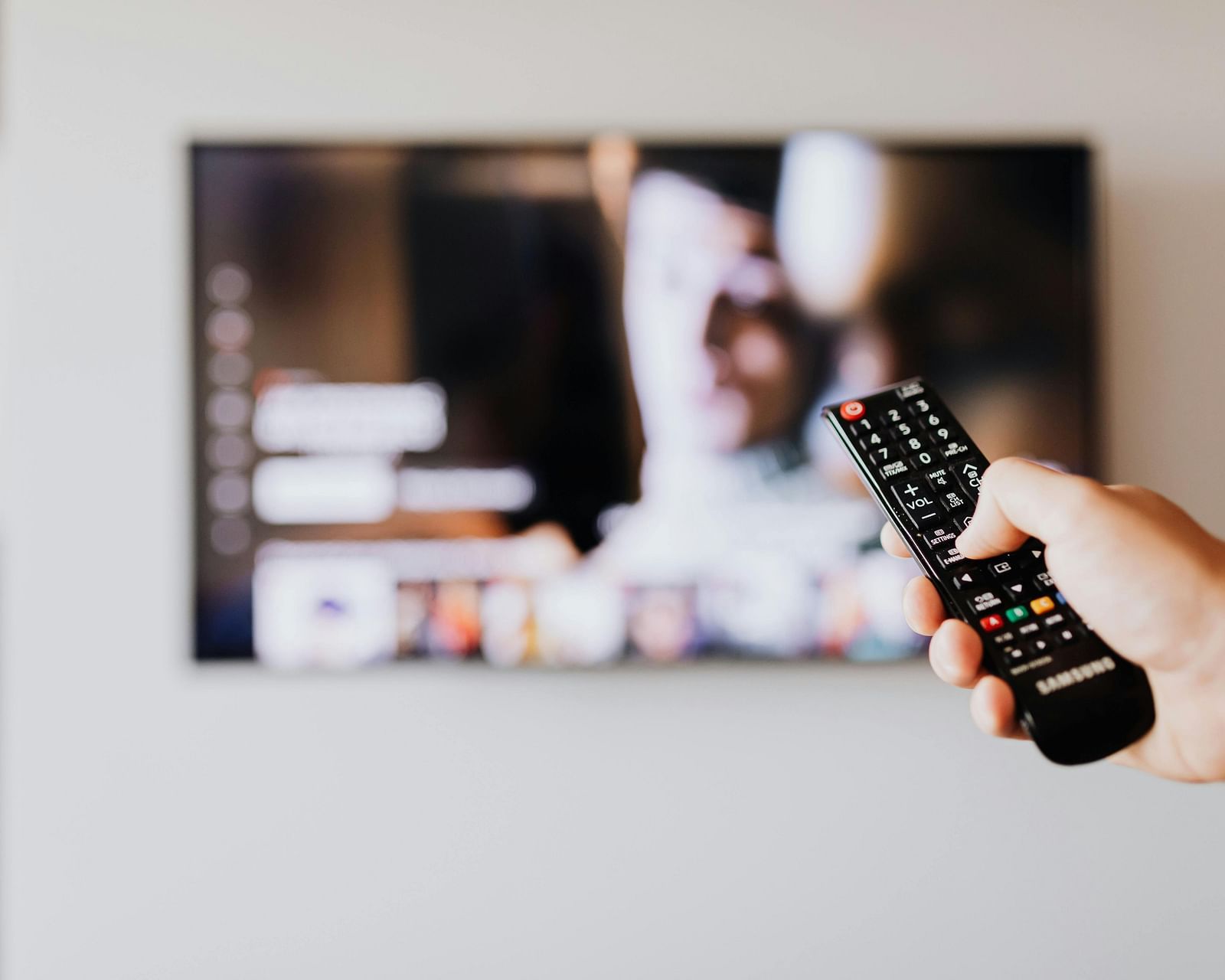 A person's hand holding a remote control pointed towards a wall-mounted television displaying content.