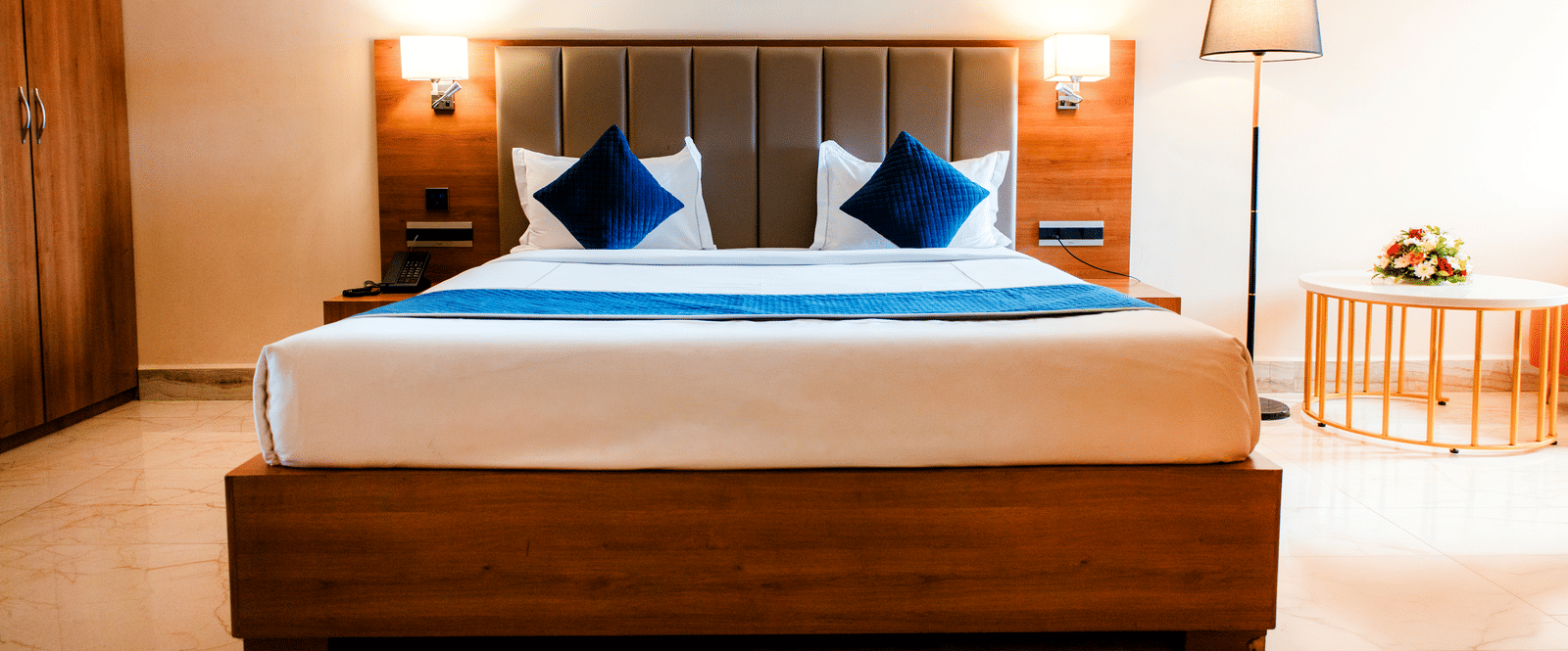 King bed in the Junior Suite with a padded headboard, teal cushions, quilted runner and bedside wall lamps at Ilara Hotels & Spa, Tada.