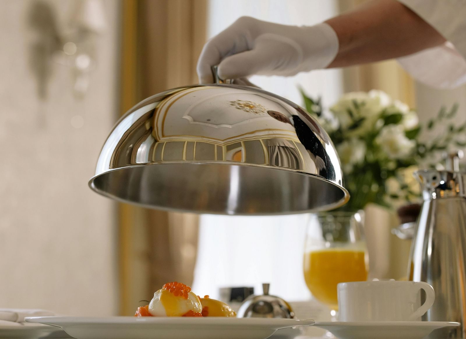 A server in white gloves lifts a silver lid to reveal a breakfast dish, with orange juice and coffee nearby.