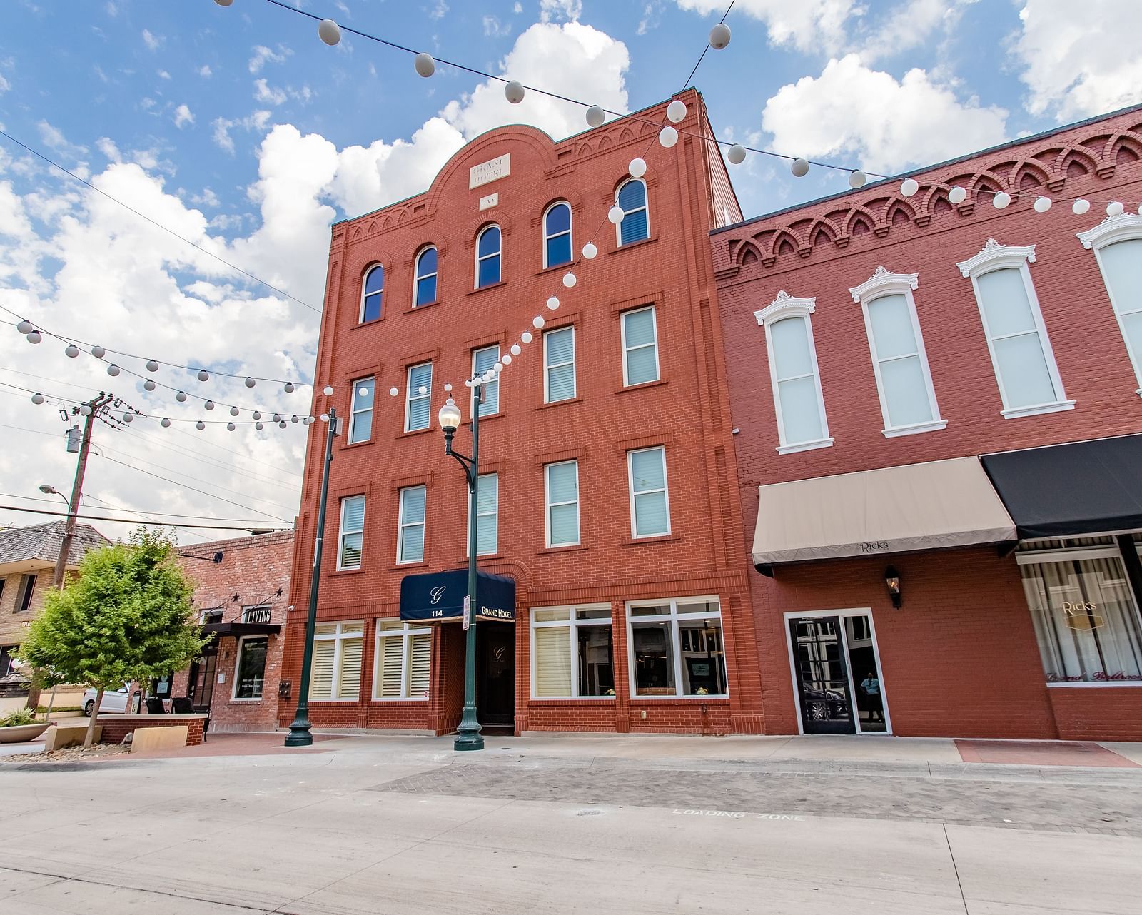 A multi-story brick building with large windows, awnings, and string lights hanging above the street.- The Grand Hotel & Ballroom, McKinney